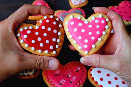 Red and pink dotted heart shaped cookies in couple's hands put together on heap of colorful cookiesの写真素材