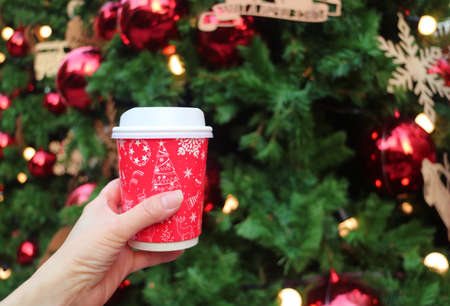 Hand Holding a Holiday Theme Takeaway Coffee Cup with Blurry Christmas Tree in Backgroundの写真素材