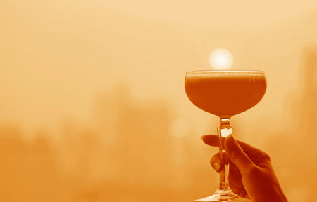 Surreal pop art style of hand holding a cocktail glass against sunset sky in soft orange color toneの写真素材