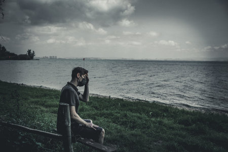A masked guy is sitting in pain on a bench on the beach near the lake with a sad, depressed, stressed expressionの写真素材