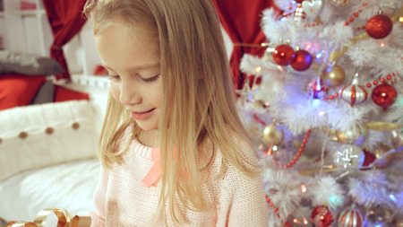 Blue-eyed little blonde girl is delighted from her New Year's gift wrapped in red paperの写真素材