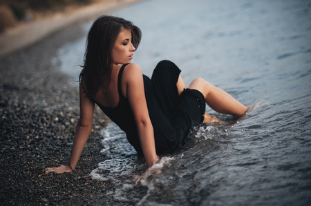 Beautiful girl wearing the black dress lying on the pebbles on the beach in the wavesの写真素材