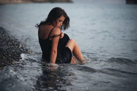 Beautiful girl wearing the black dress lying on the pebbles on the beach in the wavesの写真素材