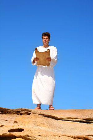 A man in a white robe reads from a scroll in a hot and sunny rocky desert landscapeの写真素材