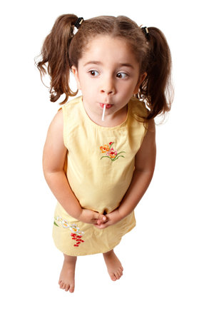 A small cute girl with hair in ponytails is sucking on a lollipop sweet.   She is standing on a white background and looking sideways.  Space for copy.の写真素材