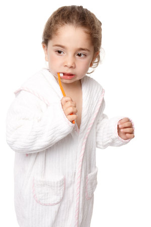 A small preschool age child brushing her teeth with an orange coloured toothbrush.  She is wearing a white bathrobe edged with pink.の写真素材