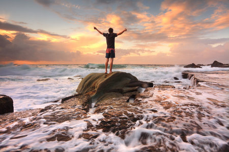 Teen boy stands on a rock among turbulent ocean seas and fast flowing water at sunrise   Worship, praise, zest, adenture, solitude, finding peace among lifes turbulent times   Overcoming adversity   Motion in waterの写真素材