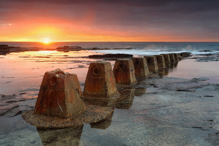 The sun rises at Coledale, casting its warm colours over the rockshelf with weathered and textured old pipeline holders in foreground.  There is a ship on the horizon.の写真素材