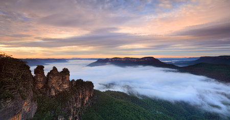 Fog filled Jamison Valley resembles a fluffy white water stream. Warm light from the sky bathes the landscape in a soft muted light and interesting clouds hand overhead.の写真素材