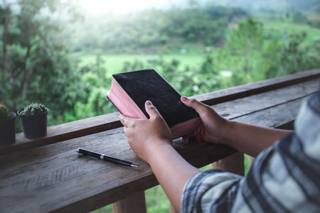 close up hands of man holding and praying with a holy bible in morning on wooden table. christian...