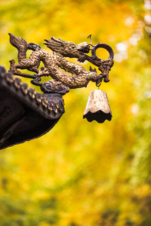 Dragon sculpture with a bell on a roof of a chinese temple with yellow leaves from autumn trees in...