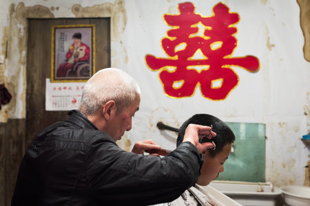 GaoMao Ancient village, Meishan, Sichuan Province, China - Apr 29, 2017: Man doing an haircut to a young boy in an old barbershop with the chinese "married" symbol on the wall.のeditorial素材