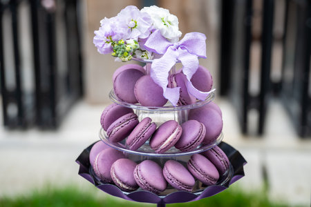 A three-tiered stand holds purple macarons with white and purple floral decorations, set against a blurred outdoor background.の写真素材