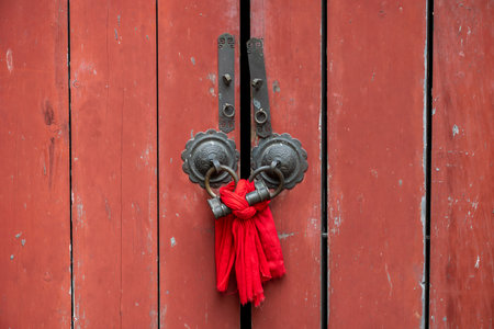 A red wooden door with a weathered texture, emphasizing its rustic charm. The door is adorned with two ornate metal hinges, each with intricate circular designs. A bright red tassel hangs in the center, adding a touch of color and cultural significance. The doors red hue contrasts with the aged wood, creating a visually striking composition.の写真素材