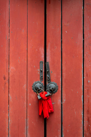 A red wooden door with a weathered texture, emphasizing its rustic charm. The door is adorned with two ornate metal hinges, each with intricate circular designs. A bright red tassel hangs in the center, adding a touch of color and cultural significance. The doors red hue contrasts with the aged wood, creating a visually striking composition.の写真素材