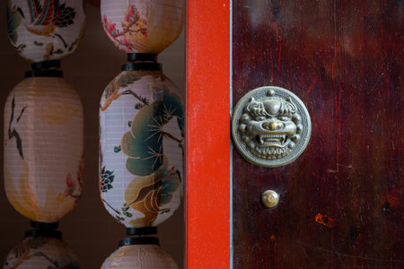A traditional Chinese door with a metallic lion-shaped door knocker. The door is made of dark wood with visible grain patterns and a red frame. To the left of the door, there are several colorful lanterns with intricate floral designs, including birds and leaves. The lanterns are arranged vertically, creating a harmonious and visually appealing composition. The dominant colors are red, brown, and pastel shades, with green and yellow floral accents.の写真素材