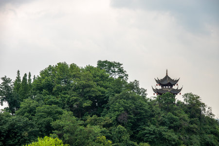 This captivating view reveals a traditional pagoda perched atop a hill, surrounded by vibrant green trees in DuJiangYan, Sichuan province. The structures intricate design features multiple eaves and decorative elements, exemplifying classic Chinese architectural style. The cloudy sky overhead adds a dramatic touch to the landscape, creating a serene atmosphere.の写真素材