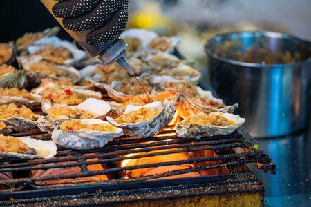 A local cook prepares fresh oysters on a bustling street in Chengdu, Sichuan. These oysters are placed on a grill, perfectly heating over bright flames. The cook skillfully adds a spicy topping, enhancing the flavors and enticing customers nearby. The atmosphere is lively with street vendors and curious onlookers enjoying the delicious aroma. This traditional dish showcases the vibrant culinary culture of Chengdu, where seafood is a popular choice among locals and tourists alike.の写真素材