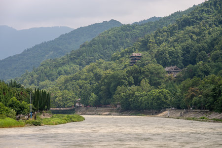 The setting features the scenic landscape of Dujiangyan in Sichuan province, where the river flows gently against a backdrop of lush mountains. The verdant hills rise steeply on either side, with trees and greenery dominating the scene. In the distance, traditional architecture peeks through the foliage, hinting at the historical importance of the area.の写真素材