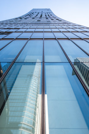 A sleek skyscraper rises dramatically against a clear blue sky. The glass facade reflects other buildings, showcasing modern architecture in a vibrant urban setting.の写真素材