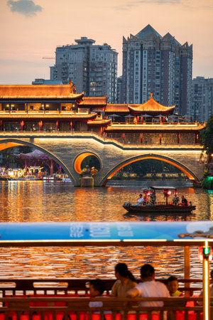 People gather near the river as boats glide through the water. The sun sets behind the bridge and buildings in Chengdu, creating a lively evening scene.のeditorial素材
