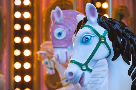 A white carousel horse with a black mane and blue eyes, wearing a green bridle, is in sharp focus against a blurred background of a pink and gold carousel with warm lights. The angle highlights the horses head, emphasizing its details and the vibrant colors of the setting.の写真素材