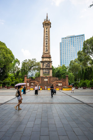 Chengdu, Sichuan province, China - Aug 20, 2025: People walk near a tall memorial monument in Peoples park. Some hold umbrellas to shield themselves from the sun. Modern buildings stand in the background among green trees.のeditorial素材