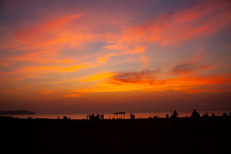 People at the beach infront of a beautiful backdrop of water and a sunset sky with yellow, red and orange colorsの写真素材