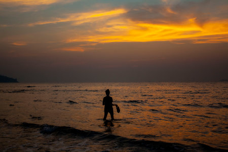 A silhouette of a girl in front of the sunset wadding in water in Goa, India. The clouds are lit a yellow and shinning of the water.の写真素材