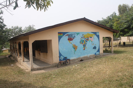 A rural school building with a colorful world map mural painted on its exterior wall. A bicycle rests against the building, adding to the scene of everyday life in a Ghanaian villageの写真素材