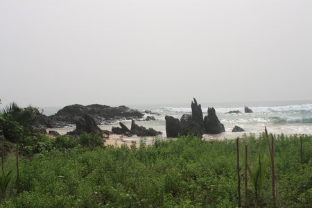 A rocky beach in Ghana with unique rock formations rising from the sand, surrounded by vibrant green vegetation, creating a striking contrast against the blue waves of the ocean.の写真素材