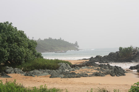 Serene beach with rocky shoreline, lush green vegetation, and a lighthouse in the distance, capturing the natural beauty of Ghana's coast.の写真素材