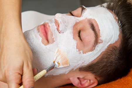 Therapist applying a face mask to a beautiful young man in a spa using a cosmetics brush の写真素材