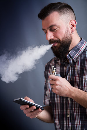 beared man  smoking electronic cigarette looking to the smartphoneの写真素材