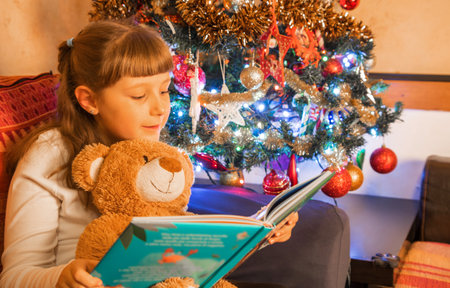 young girl reads a book in front of the Christmas tree with her teddy bearの写真素材