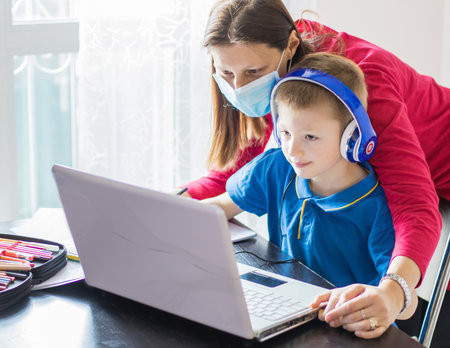 Coronavirus Outbreak. Lockdown and school closures. Mother helping her son with face mask studying online classes at home. COVID-19 pandemic forces children and teachers online learning.の写真素材