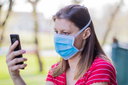 caucasian woman with mask looking out to home terrace using mobile phone during quarantine due to coronavirus covid19 pandemic.の写真素材