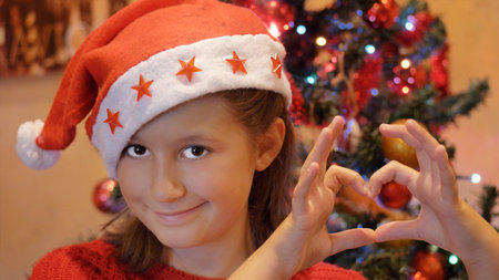 young girl with Sants Claus Hat doing heart shape with hands with christmas tree as background.の写真素材