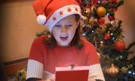 young girl with Sants Claus Hat doing heart shape with hands with christmas tree as background.の写真素材