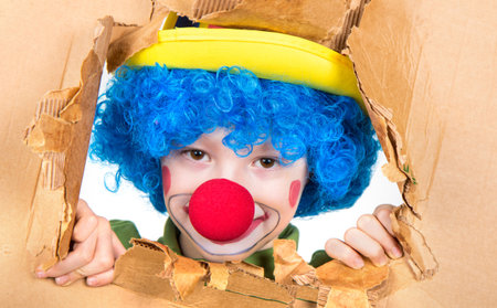 young child dressed as a clown with wig and fake nose has fun watching from the open cardboardの写真素材
