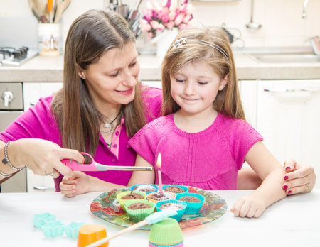 Cute little girl blowing out candles with mom on birthday cake muffin at homeの写真素材