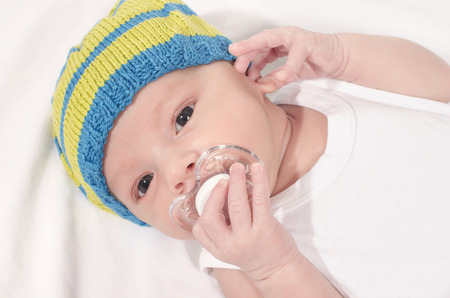 Beautiful baby lying down on white sheets with a pacifier in his mouth. Cute new born baby boy wearing a hat holding a binky.の写真素材