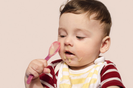 Child trying to feed himself baby food holding the spoon by himself. Six month old infant beginning food diversification.Cute baby boy wanting to eat food with the spoon.の写真素材