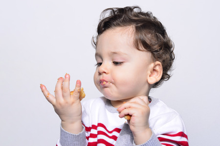 Portrait of a cute baby eating a biscuit looking at the hand.One year old kid eating biscuits by himself. Adorable curly hair boy being hungry.の写真素材