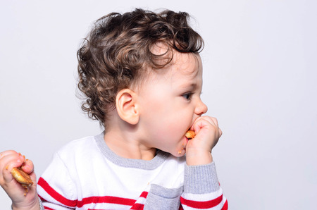 Portrait of a cute baby eating a biscuit. One year old kid eating biscuits by himself. Adorable curly hair boy being hungry.の写真素材