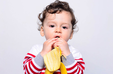 Portrait of a cute baby eating a banana. One year old kid eating fruits by himself. Adorable curly hair boy being hungry.の写真素材
