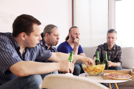 Group of men drinking beer, eating pizza, talking and smiling while resting at home on couch behind TVの写真素材