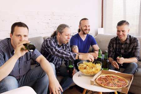 Group of men drinking beer, eating pizza, talking and smiling while resting at home on couch behind TVの写真素材