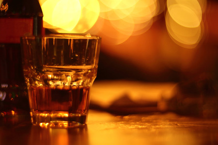 Glass of scotch whiskey and bottle on table in bar over bokeh golden glowing lights background