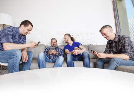 Four happy male friends using their mobile phones sitting on a sofa at homeの写真素材